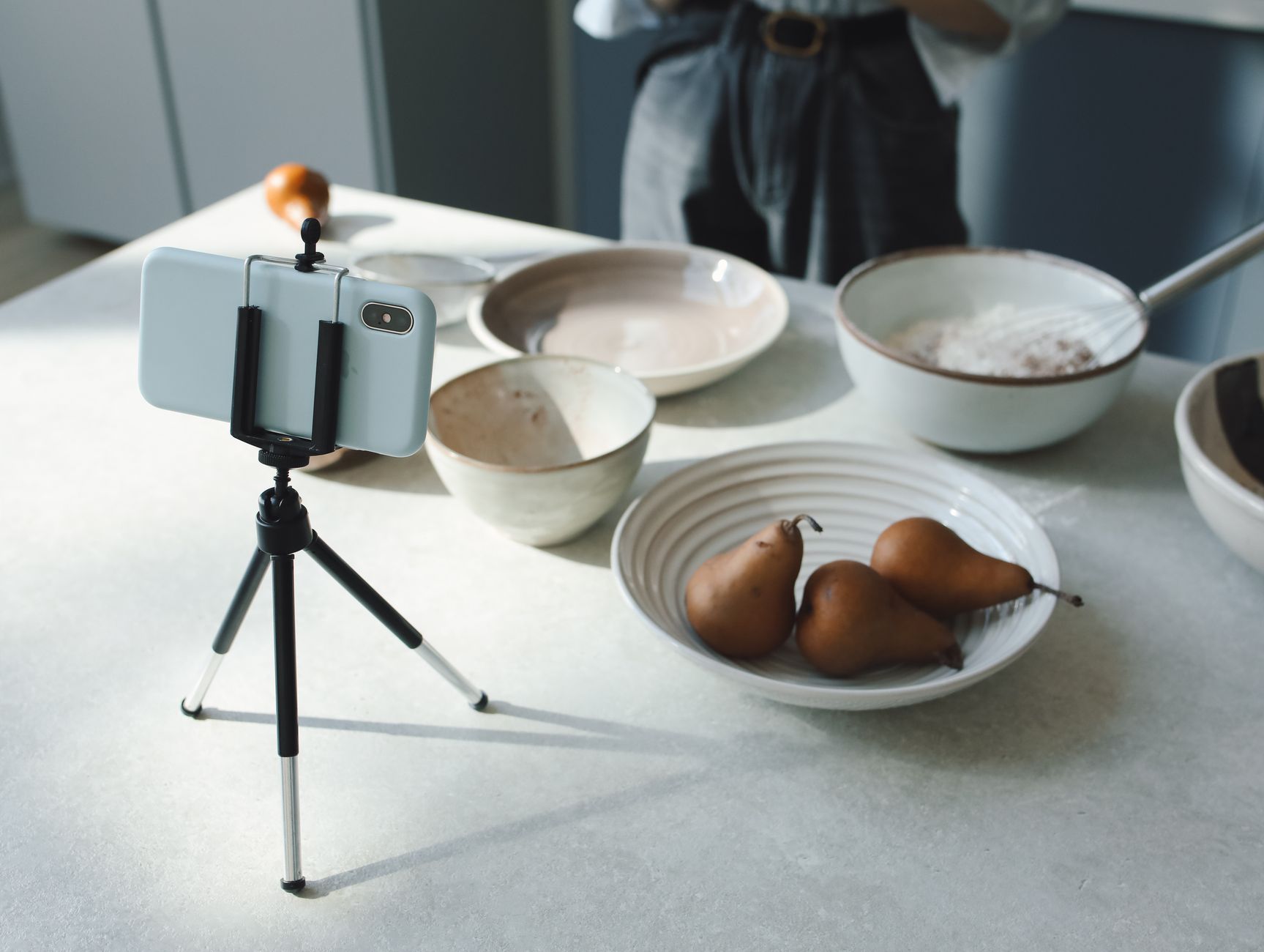 Un smartphone fixé sur un trépied de table, face à un plan de travail avec des bols en céramique et trois poires.