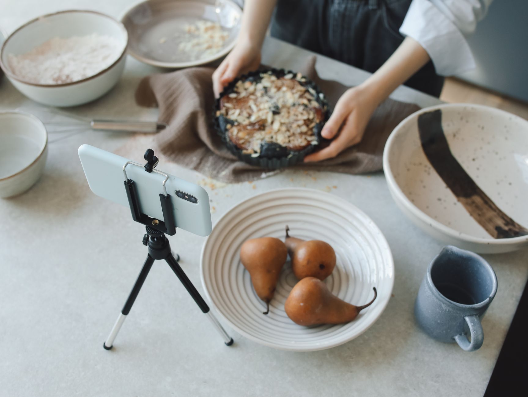 Une personne présente un gâteau aux poires dans une cuisine pendant qu’un smartphone posé sur un trépied filme la scène.