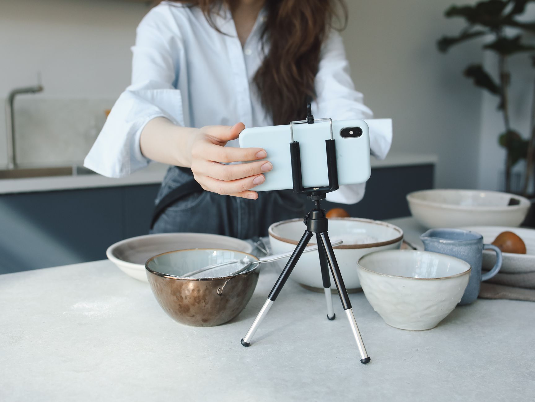 Une femme règle son smartphone fixé sur un petit trépied, posé sur un plan de travail de cuisine entouré de bols en céramique, pour filmer une vidéo.